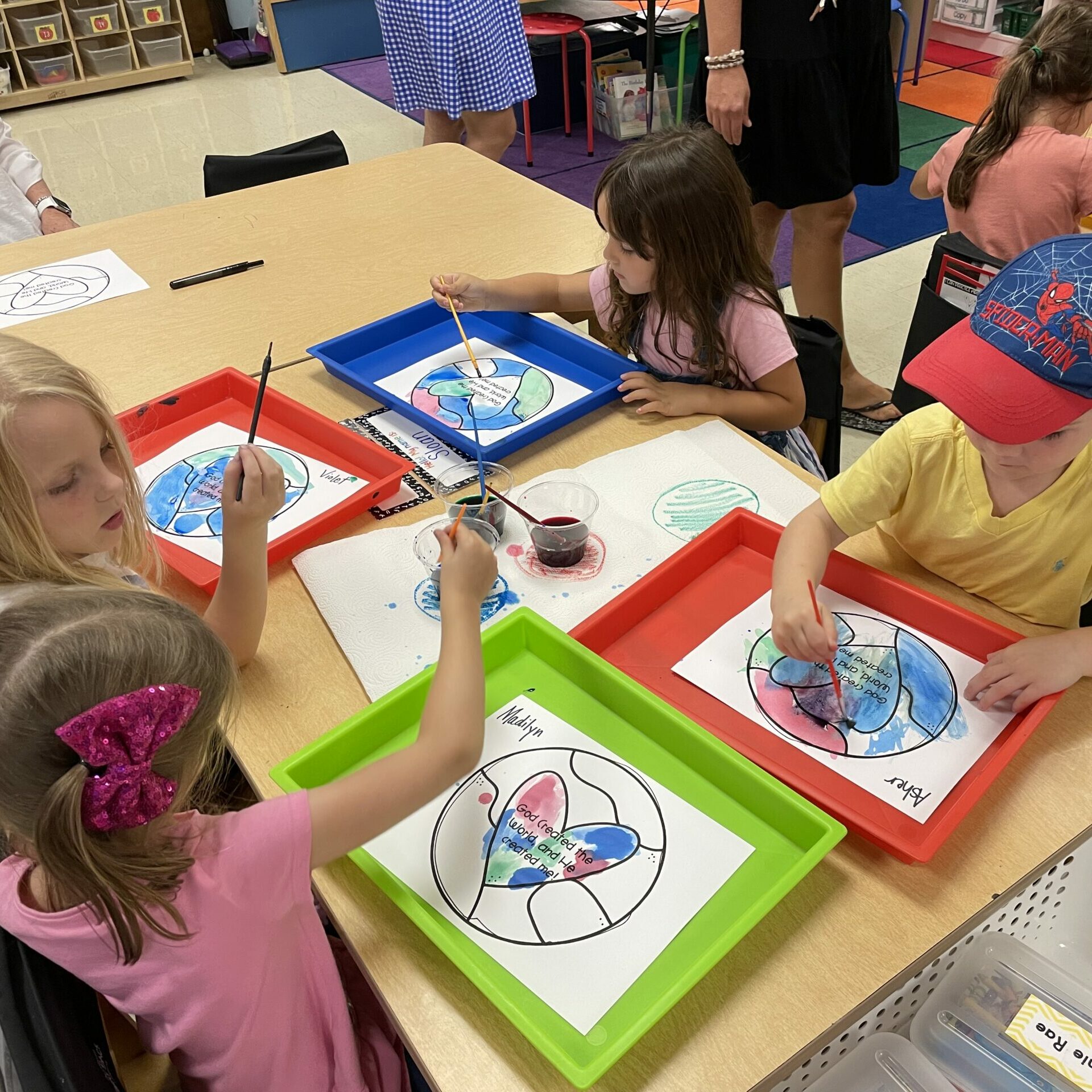 A boy child is sitting quietly in School class, coloring a picture, as teachers walk around behind him.